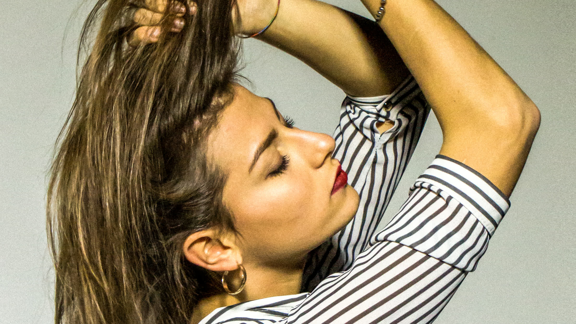 I capelli tirati su con entrambe le mani, il viso di profilo rivolto verso l&rsquo;alto, la camicia a righe