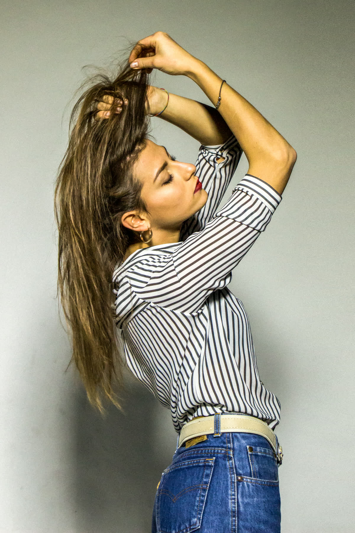 I capelli tirati su con entrambe le mani, il viso di profilo rivolto verso l&rsquo;alto, la camicia a righe